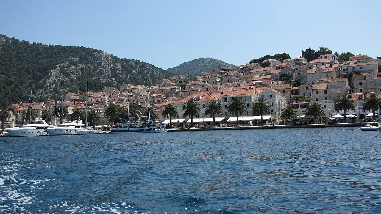 Hvar town seen from the water, stone houses and terracotta roofs climbing the hillside above a palm-lined harbour with yachts moored below