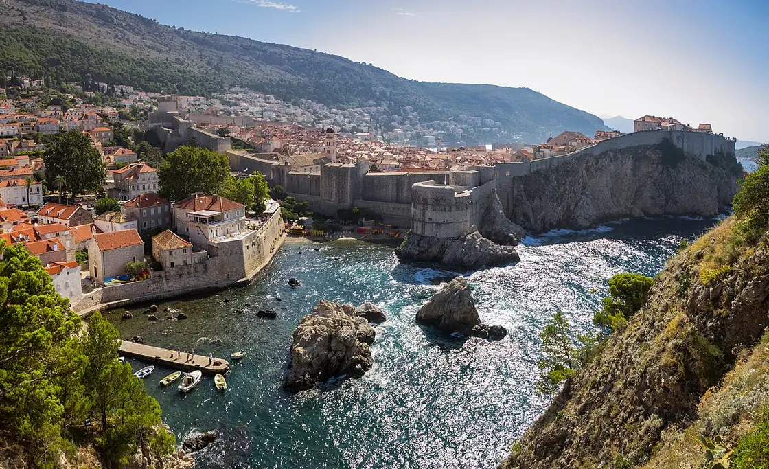 The stone city walls of Dubrovnik rising above a rocky cove on the western side, with Fort Bokar and the terracotta roofs of the old town behind