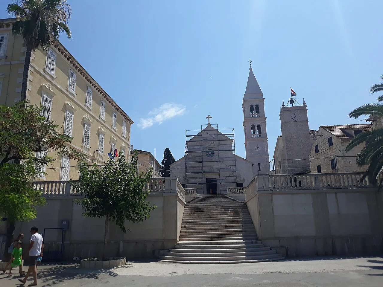 A palm-lined waterfront street in Supetar on Brač with stone houses and moored boats