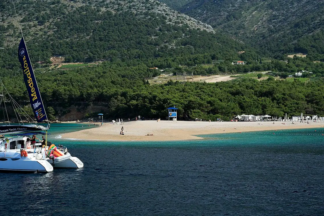 Turquoise water lapping the white pebbles and pines of Zlatni Rat with people swimming