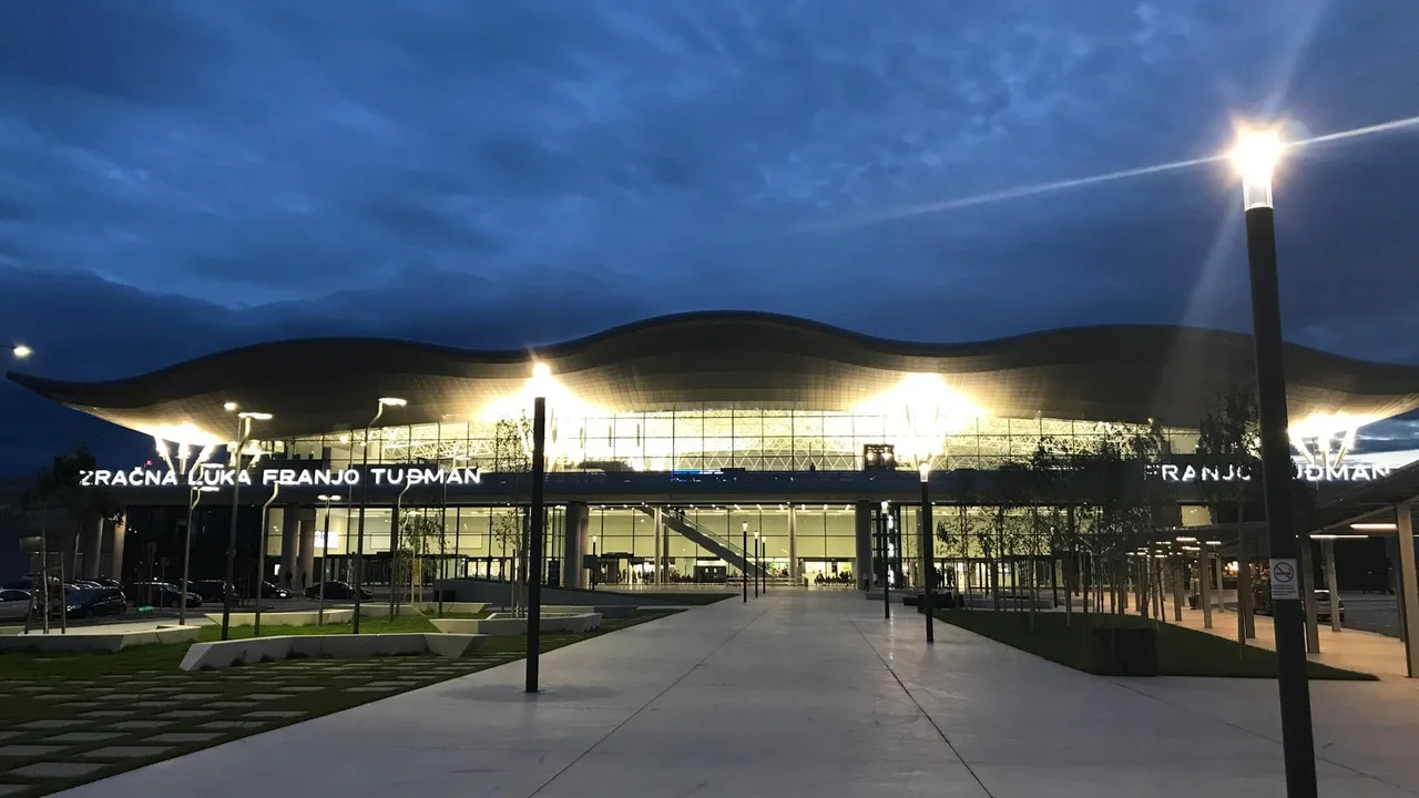 The lit glass front of the Franjo Tudjman Airport terminal in Zagreb at dusk, its wavy roof above the entrance and cars parked at the kerb