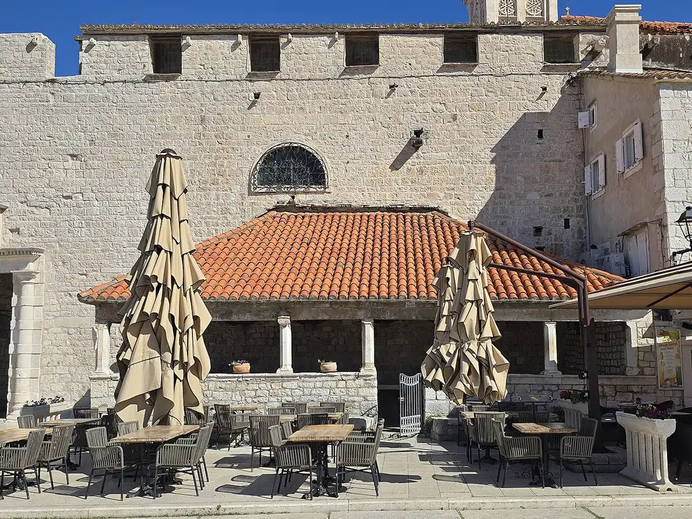 An empty cafe terrace with folded parasols in front of a stone loggia in Trogir old town