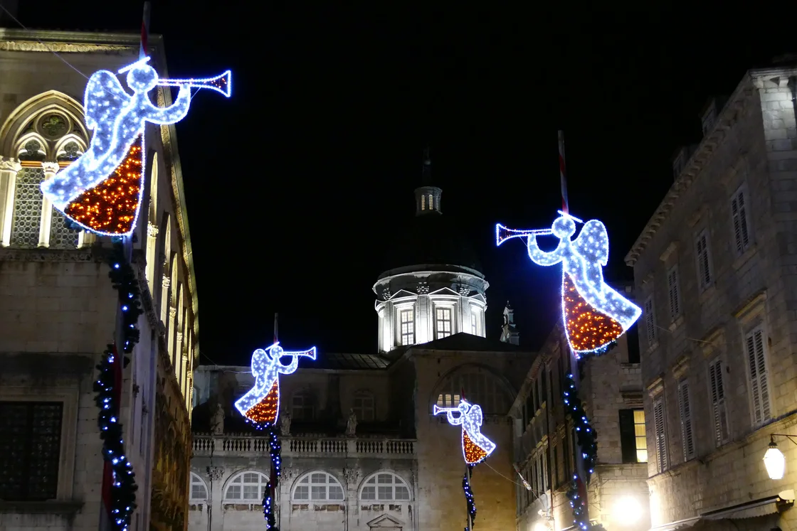 Lit angel figures with trumpets on a Dubrovnik street at night with a church dome behind