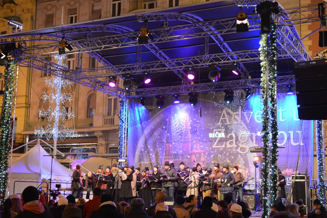 A choir on a lit stage with an Advent banner on Ban Jelačić Square in Zagreb, with a tram behind