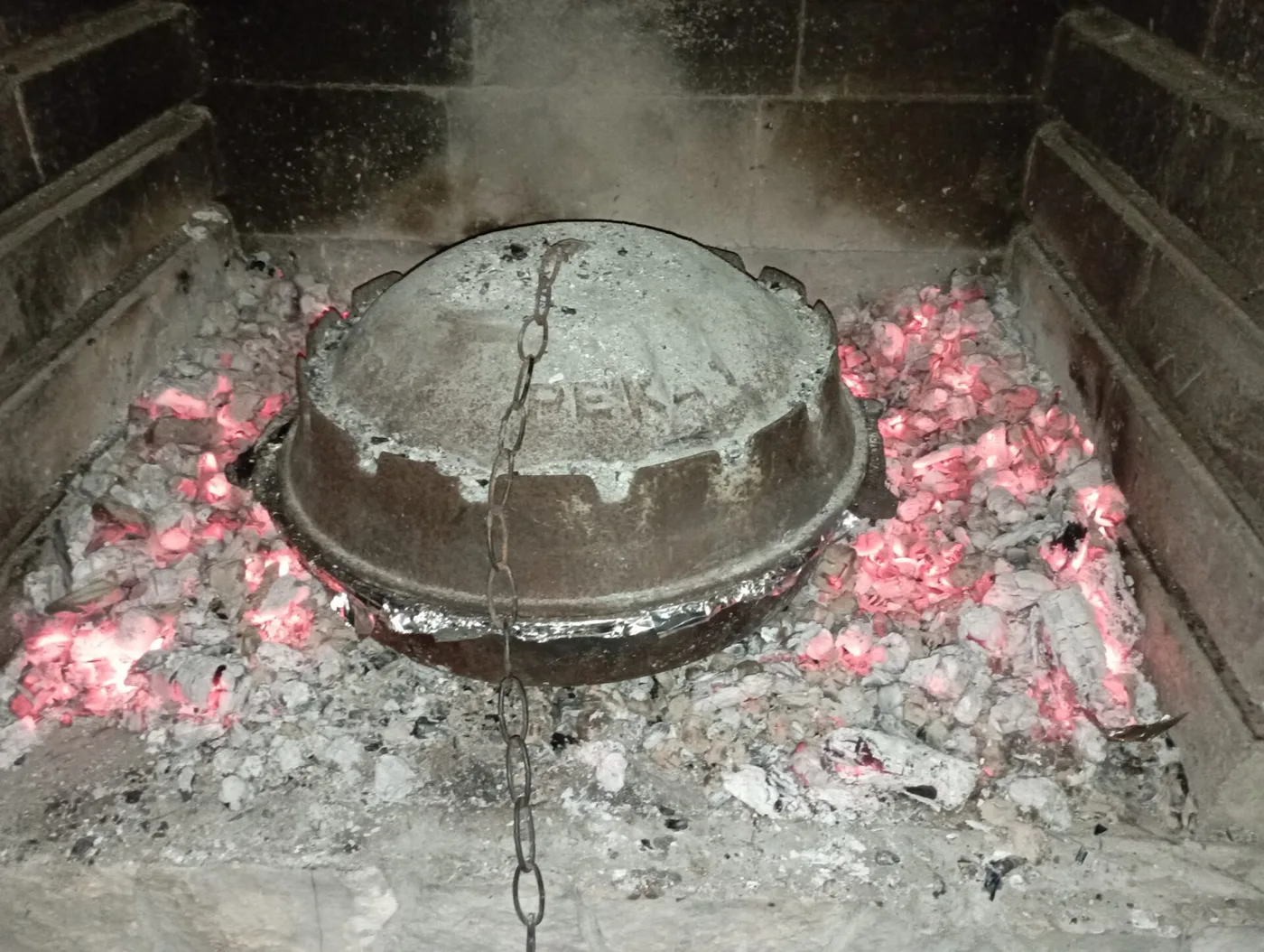 A metal peka bell lid lifted off a dish of slow-roasted meat and potatoes cooked under embers
