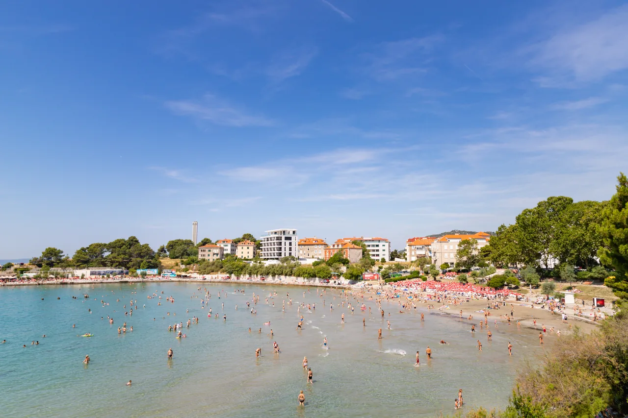 A packed sandy Croatian beach on a hot July day, with swimmers, red parasols and a bright blue sky