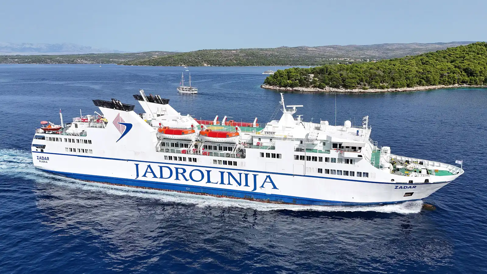 The stern of an off-season Croatian ferry near Makarska, empty yellow benches and a flag snapping in the wind, with the Biokovo mountains behind