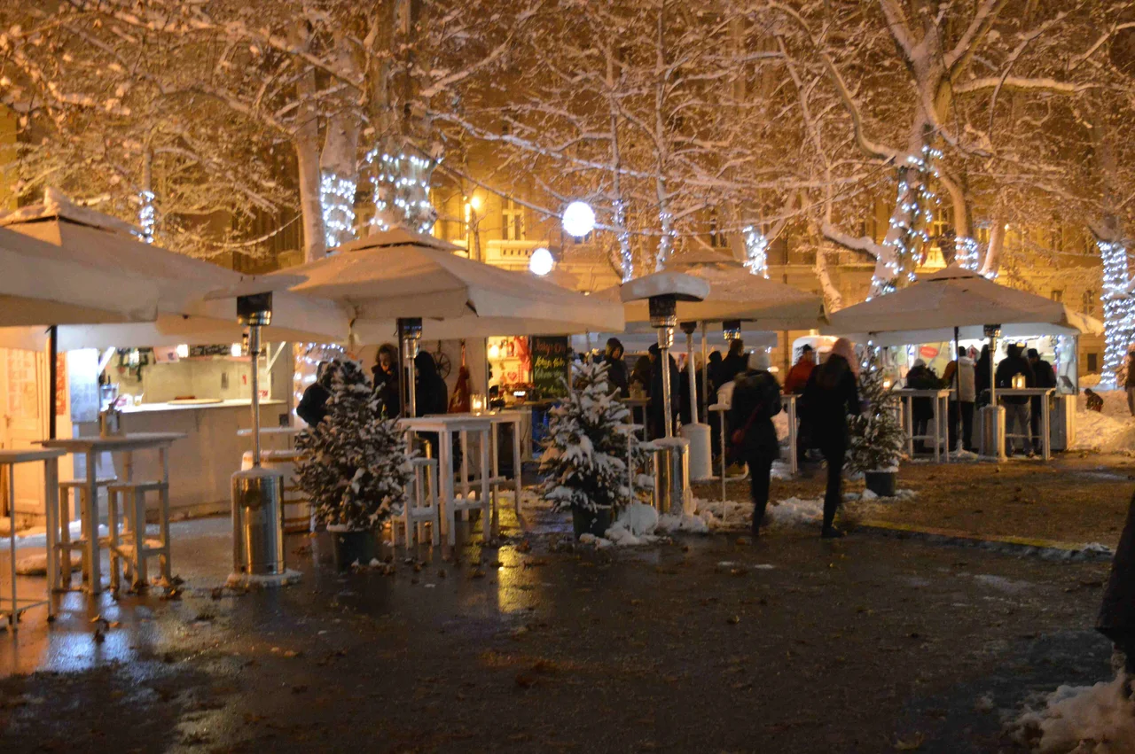 Advent huts and umbrellas dusted with snow under snow-covered plane trees wrapped in fairy lights at night, with a few visitors at the counters
