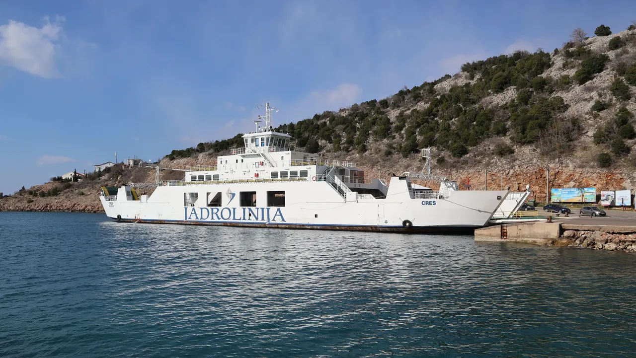 The white Jadrolinija car ferry Cres moored at a quay below a rocky hillside under a blue sky