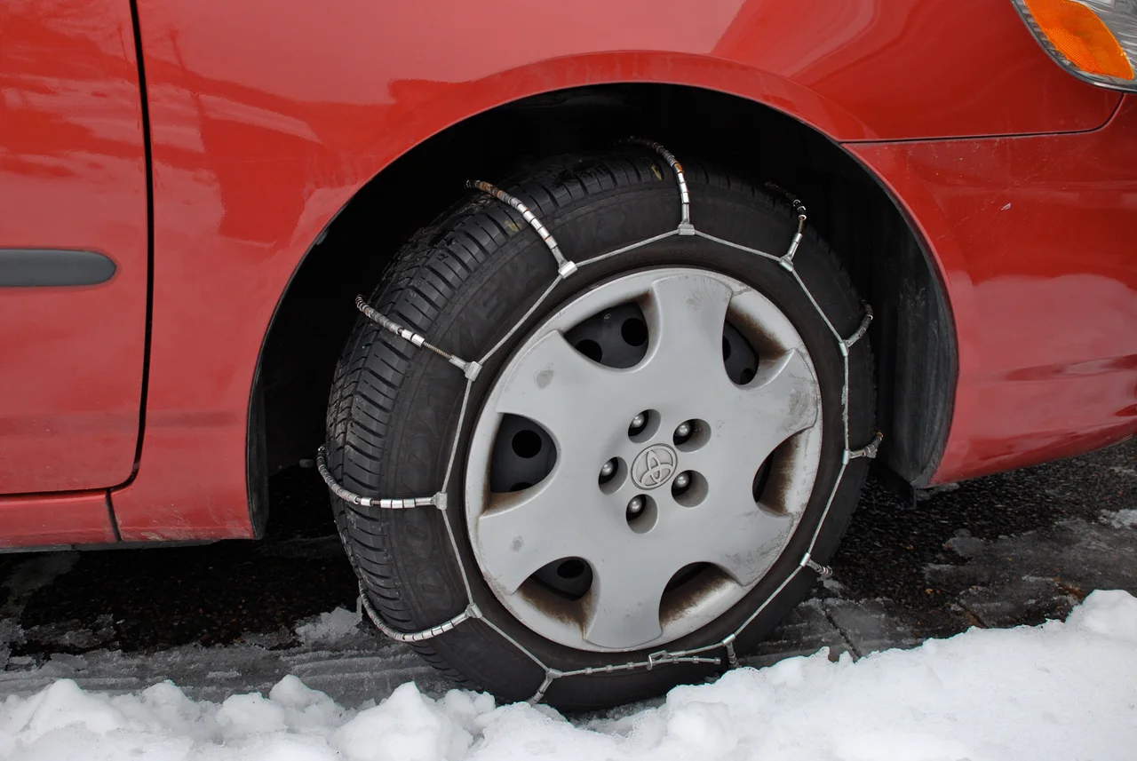 Cable-type snow chains fitted on the front wheel of a red car standing in slush