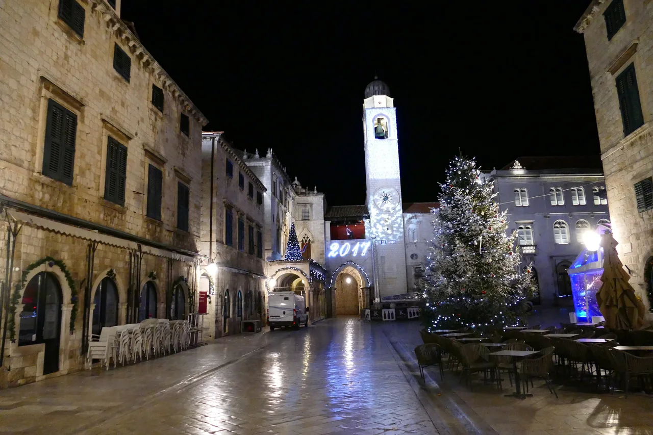 Empty Stradun in Dubrovnik at night with a lit Christmas tree, stacked cafe chairs and a floodlit bell tower
