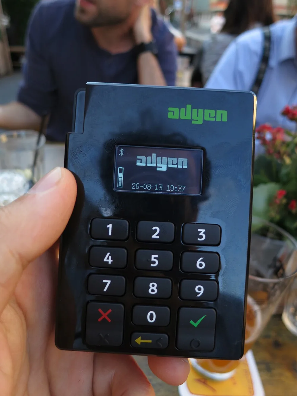 A hand holding a small card payment terminal at an outdoor café table