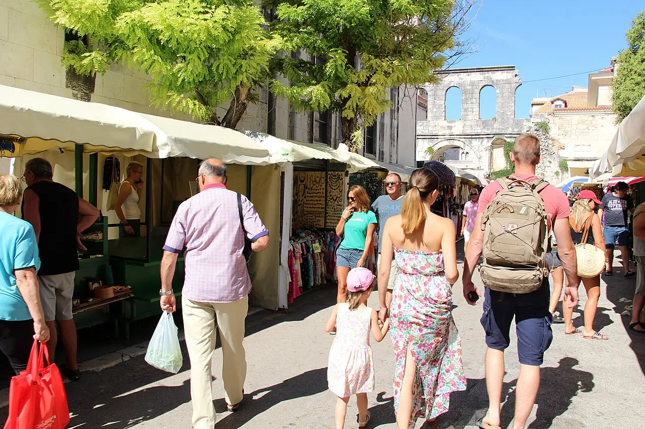 Shoppers walking through the open-air Stari pazar market in Split beside the Roman Silver Gate, stalls with clothes and goods under awnings