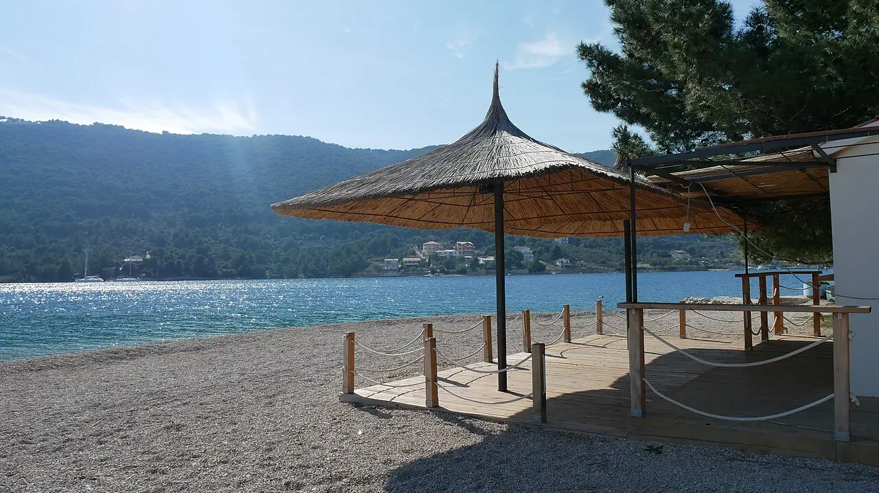 A pebble beach in Croatia with a thatched parasol, pine trees and clear blue water