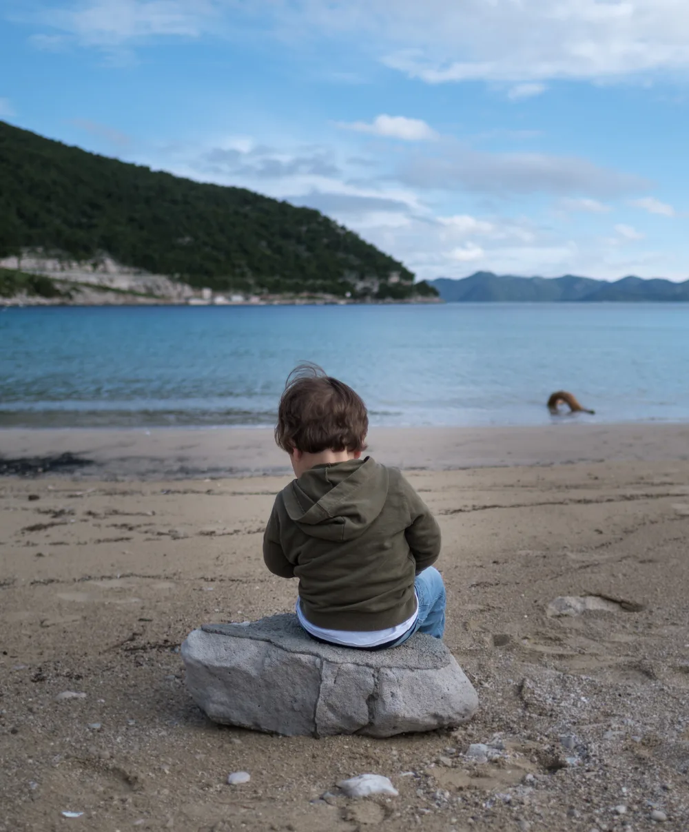 A child in a hooded top sitting on a rock on an empty sandy Adriatic beach under a bright but cool sky
