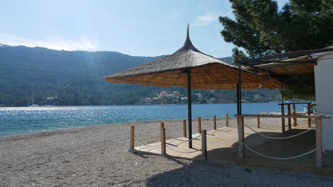 A calm bay at Grebastica with a thatched sun parasol on a pebble beach and sparkling water in morning light
