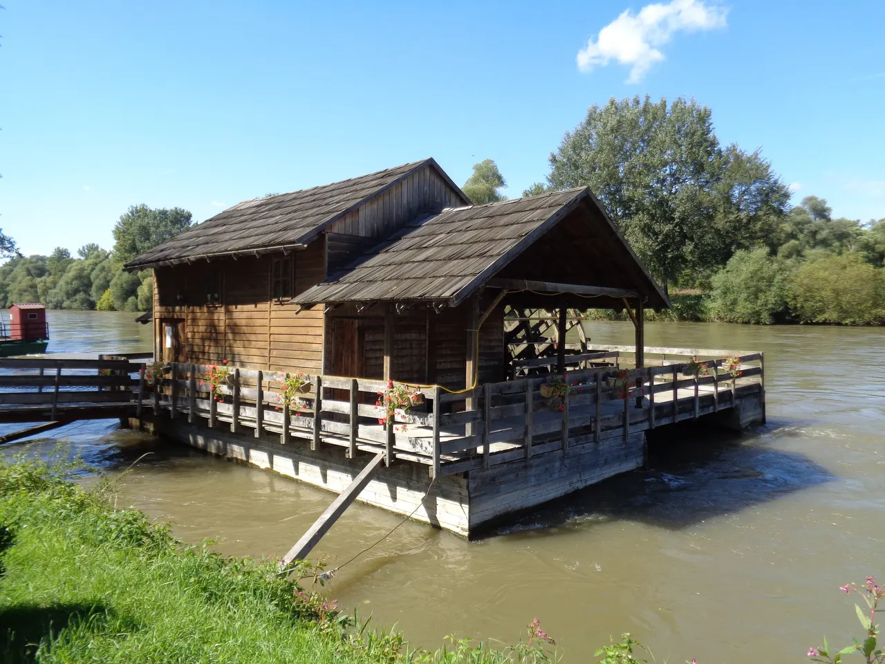 A wooden floating watermill moored on the Mura river at Sveti Martin na Muri, with a footbridge and flower boxes