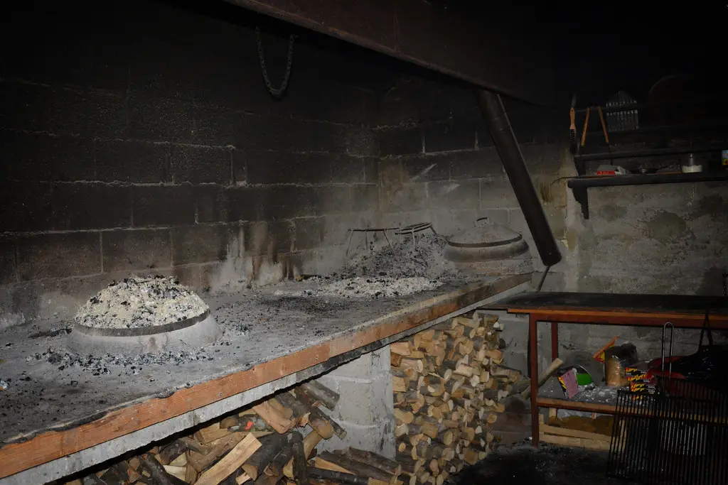Two peka lids buried under grey ash in a konoba fireplace with firewood stacked underneath