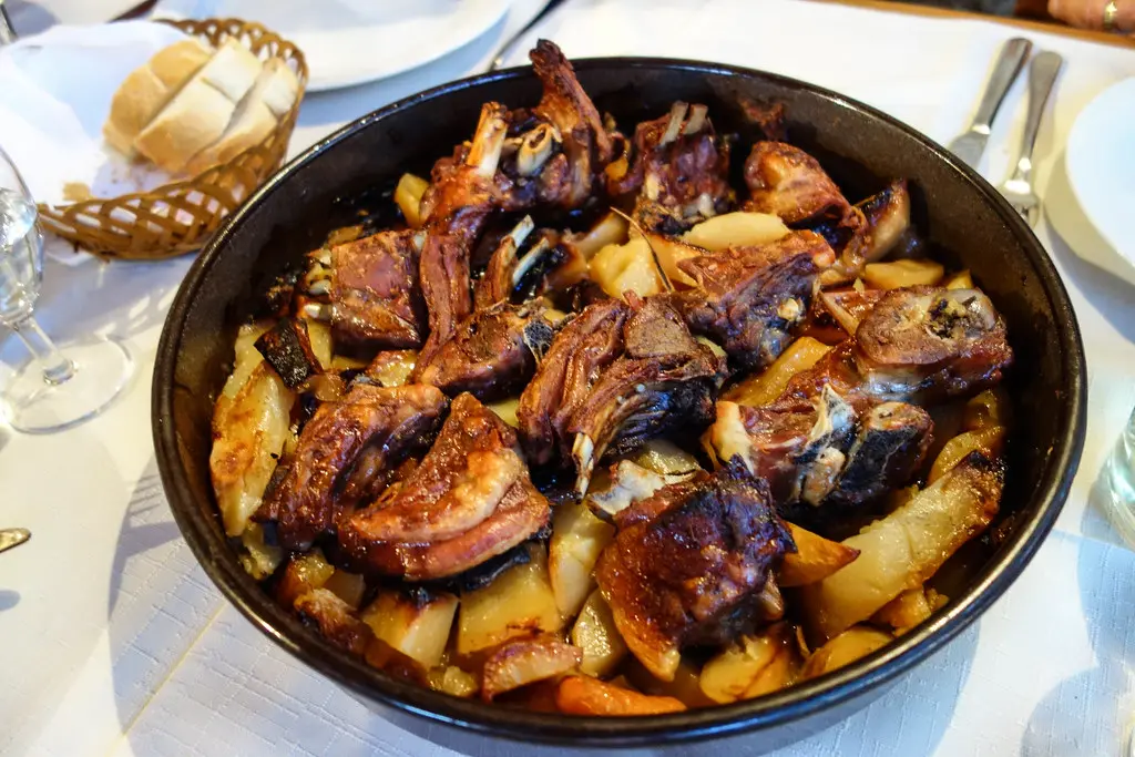 A black pan of lamb peka with browned meat and potatoes on a restaurant table with bread