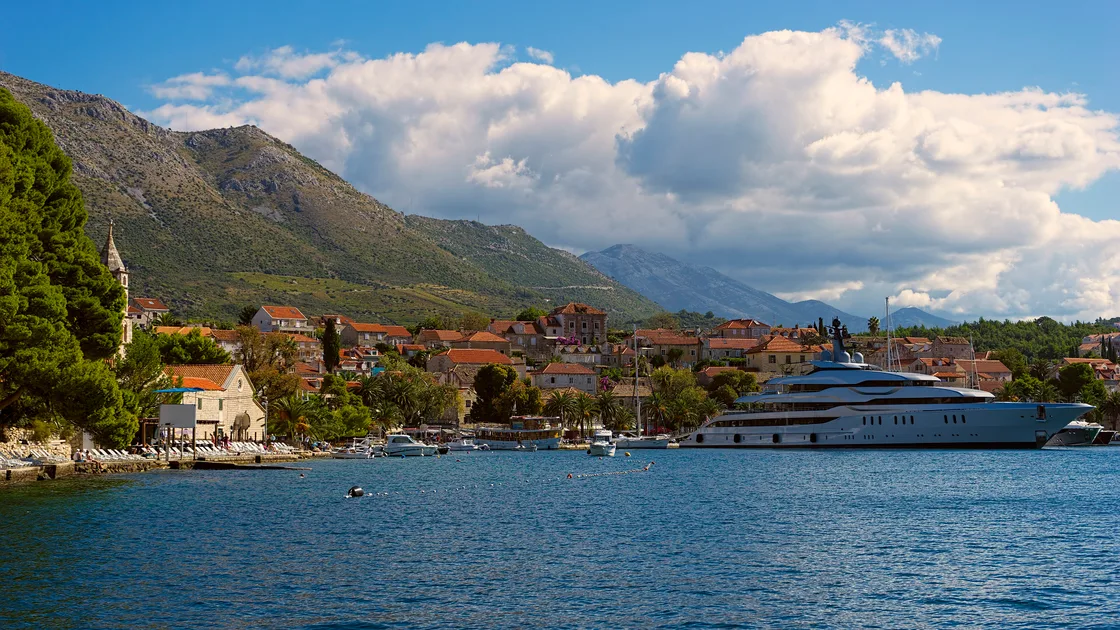 Sunlit waterfront of Cavtat with a stone church, red-roofed houses, palms and boats in the harbour below green mountains