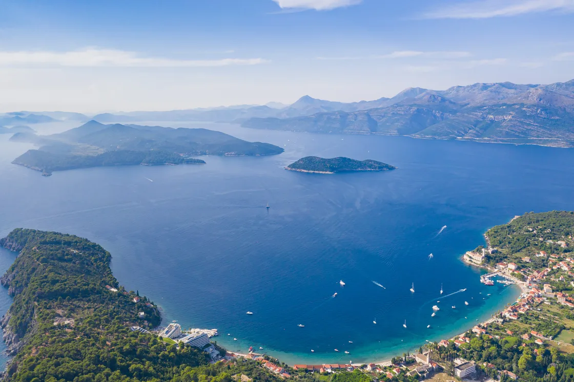 Aerial view of Lopud island in the Elaphiti archipelago, with a curved sandy bay, moored boats, a village and green hills