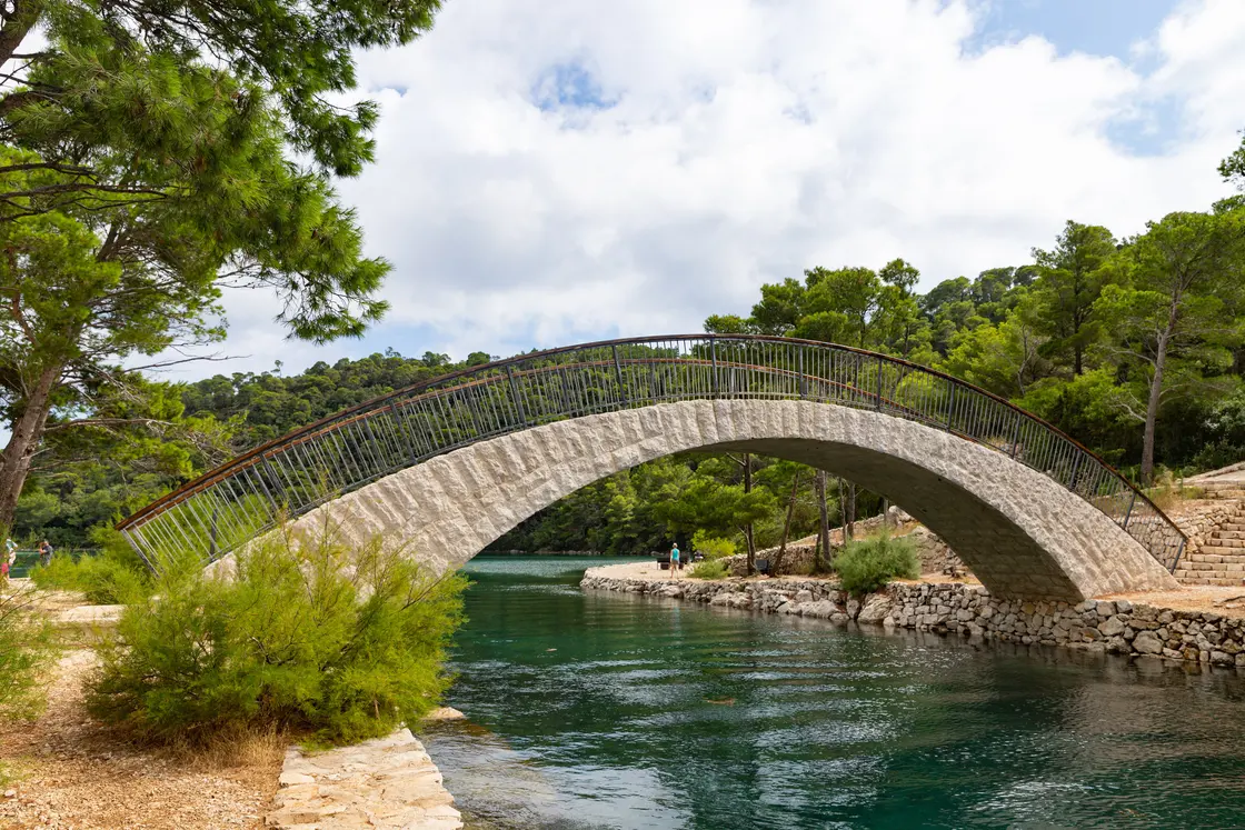 A stone arched footbridge over a narrow turquoise channel between the two lakes of Mljet, surrounded by pine forest