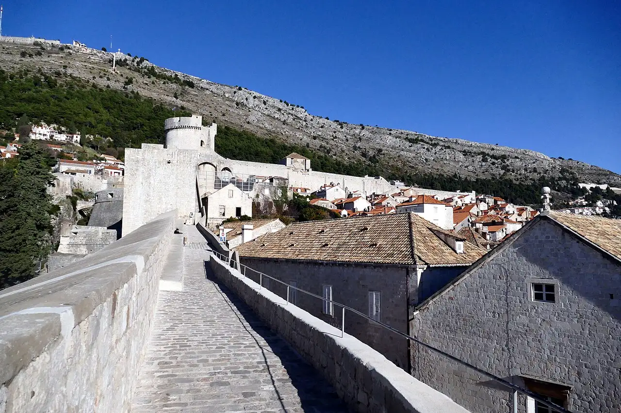 The stone walkway on top of the Dubrovnik city walls leading toward the round Minceta Tower, with Mount Srd behind