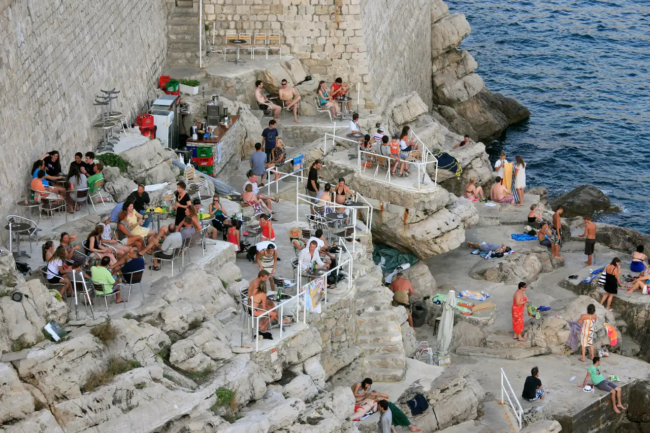 Tables and chairs spread across rock ledges below the Dubrovnik city walls, with people drinking, sunbathing and swimming at the cliff bar
