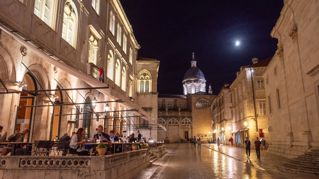 Diners at a lit terrace on a Dubrovnik Old Town street at night, with the cathedral dome and a full moon behind