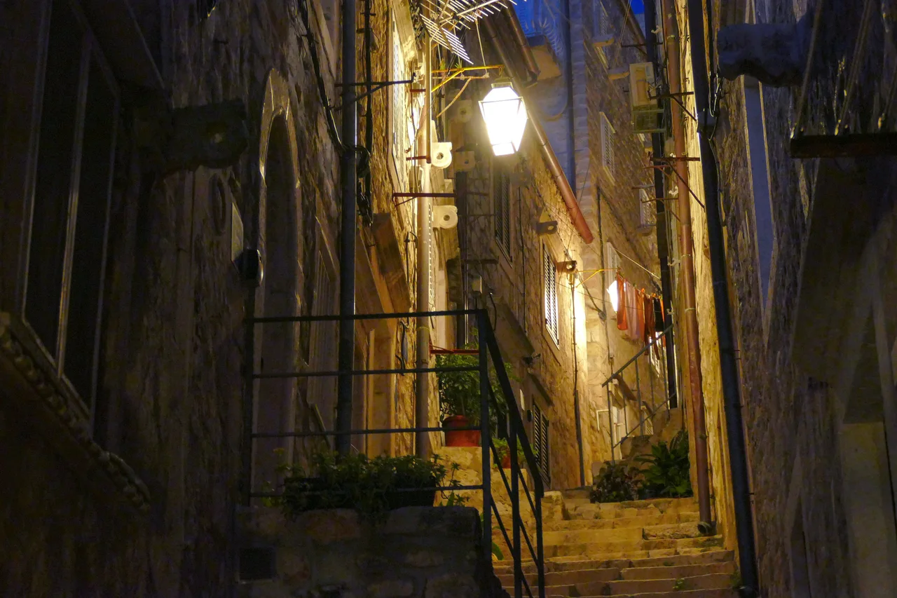 A narrow stepped lane in Dubrovnik Old Town at night, lit by a single lantern, with potted plants and washing hanging between stone houses