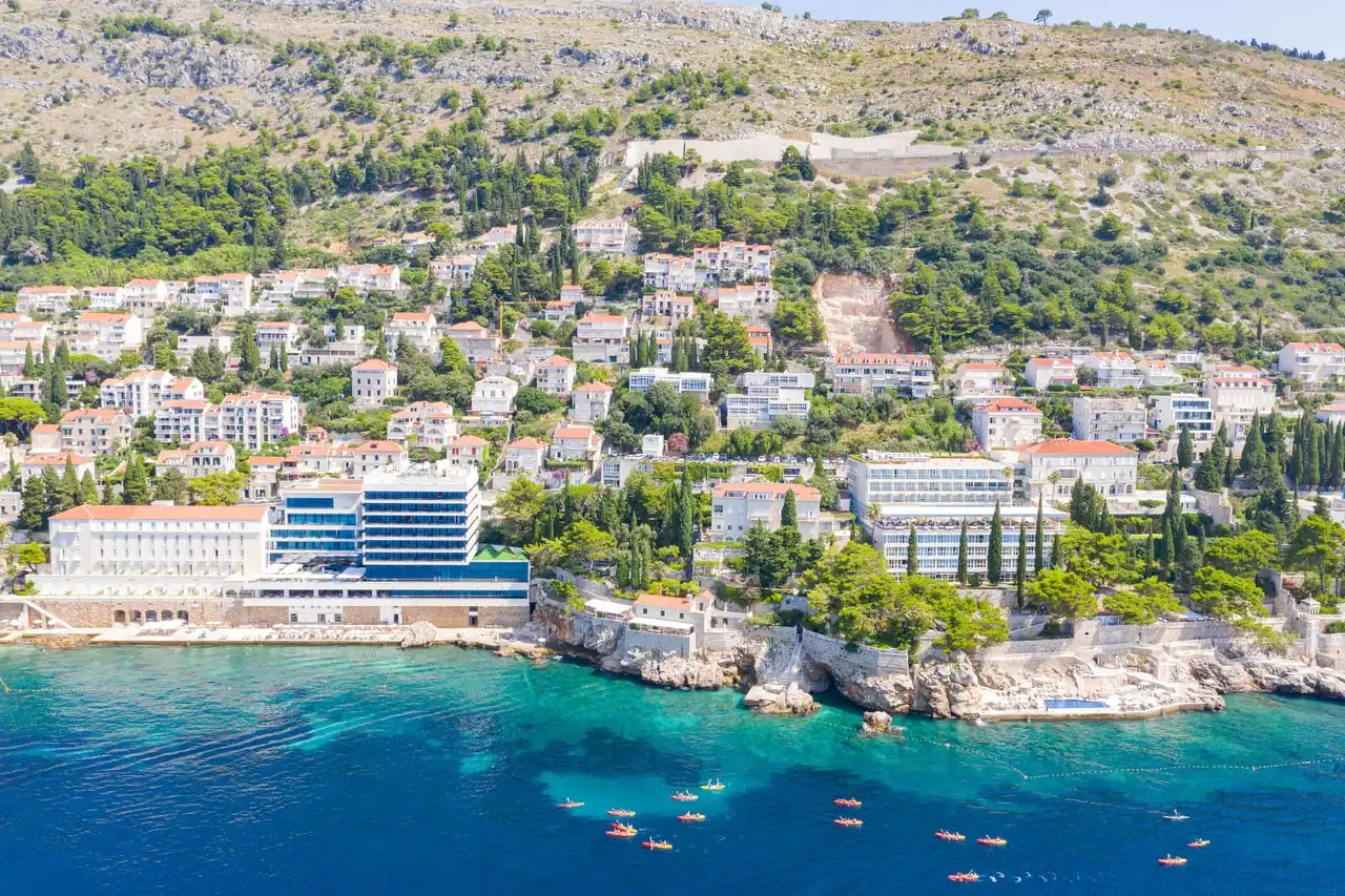 A group of sea kayaks on turquoise water below the hotel-lined coast of Dubrovnik, seen from the air