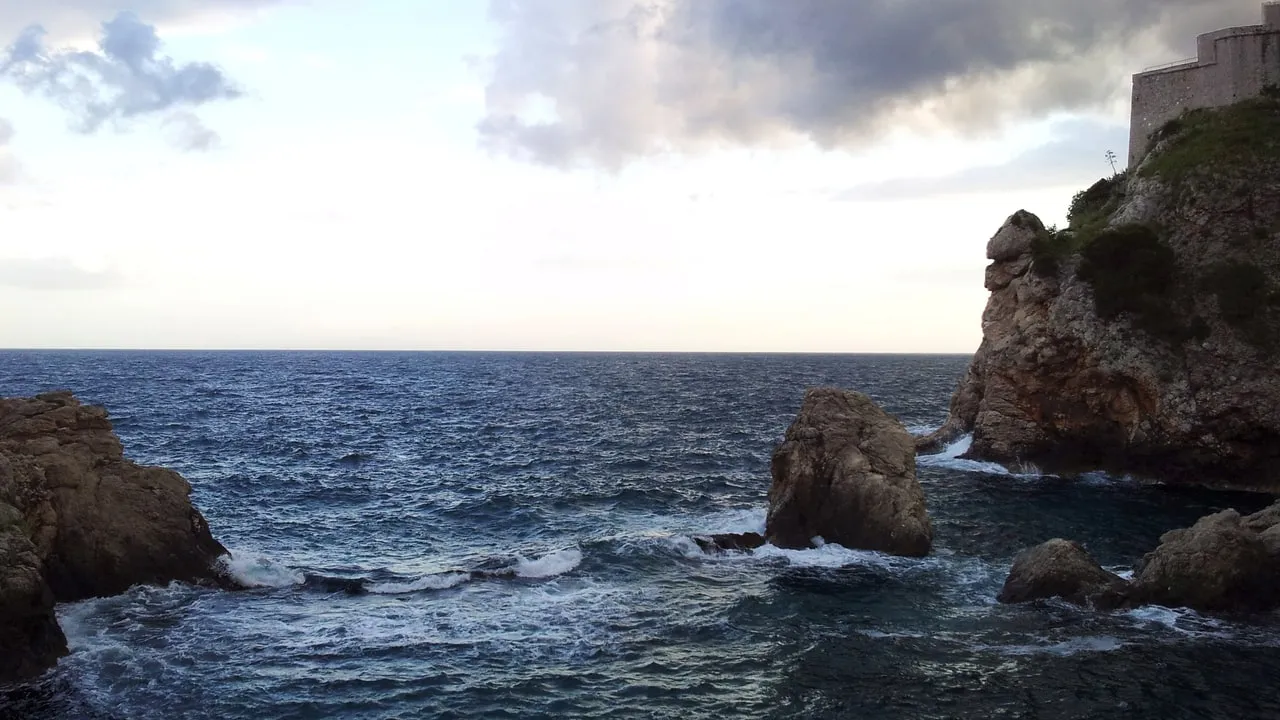 Choppy sea between rocks under a stormy sky, with the stone wall of Fort Lovrijenac at the edge of the frame