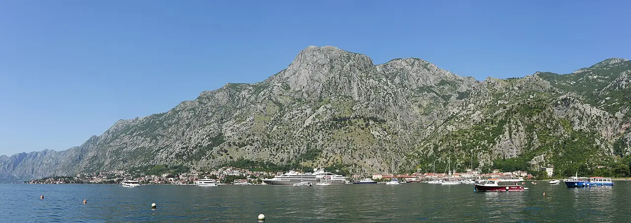 Wide panorama of the Bay of Kotor with a huge grey mountain rising straight from the water and boats moored below