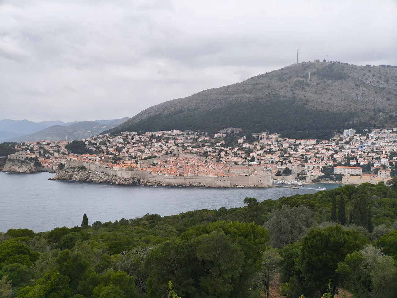 The walls and orange roofs of Dubrovnik's old town under a grey cloudy sky, seen across the water from a green hillside, with a bare mountain behind