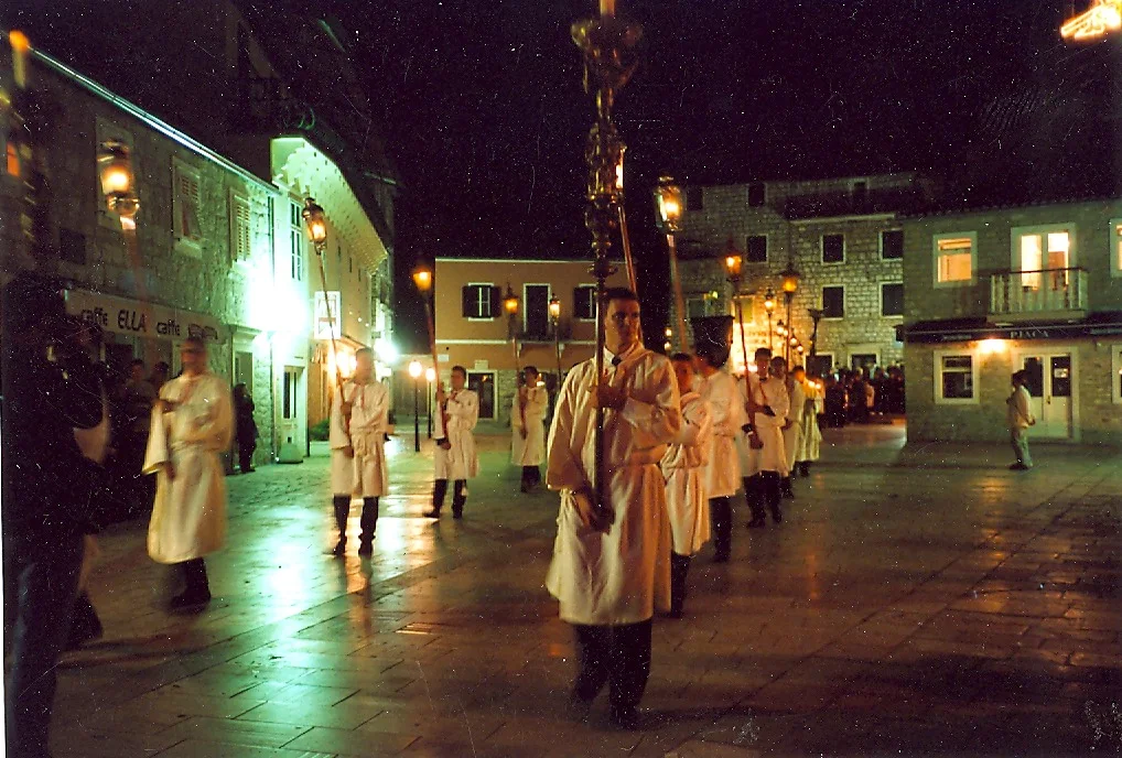 Men in white robes carry tall processional lanterns in a line across a lit stone square at night, with cafe fronts and stone houses behind them