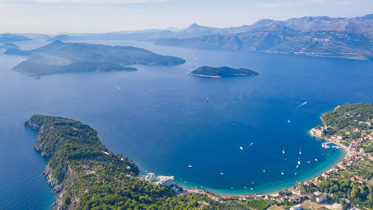 Aerial view of Lopud harbour with terracotta roofs, anchored boats and a wooded island beyond