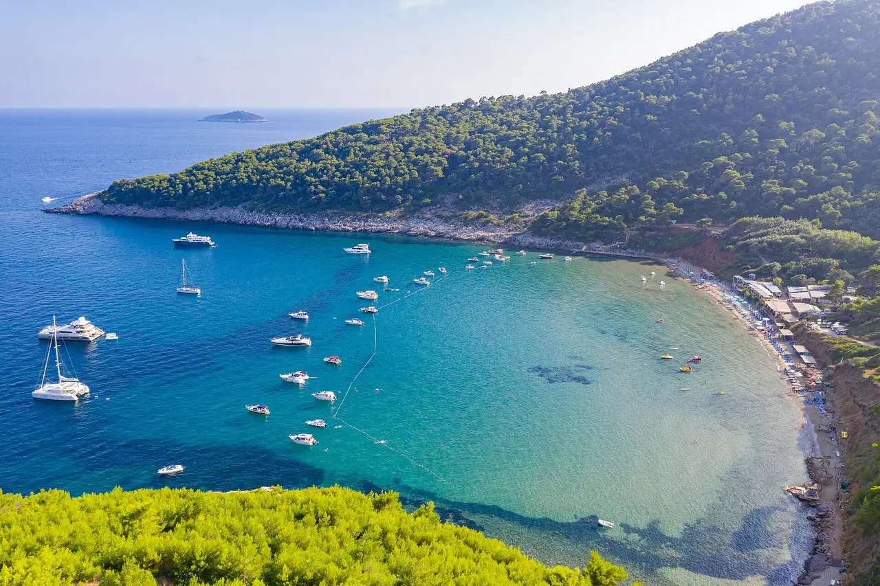 Aerial view of Šunj bay on Lopud with turquoise shallows, a buoy line and boats anchored outside the swimming area