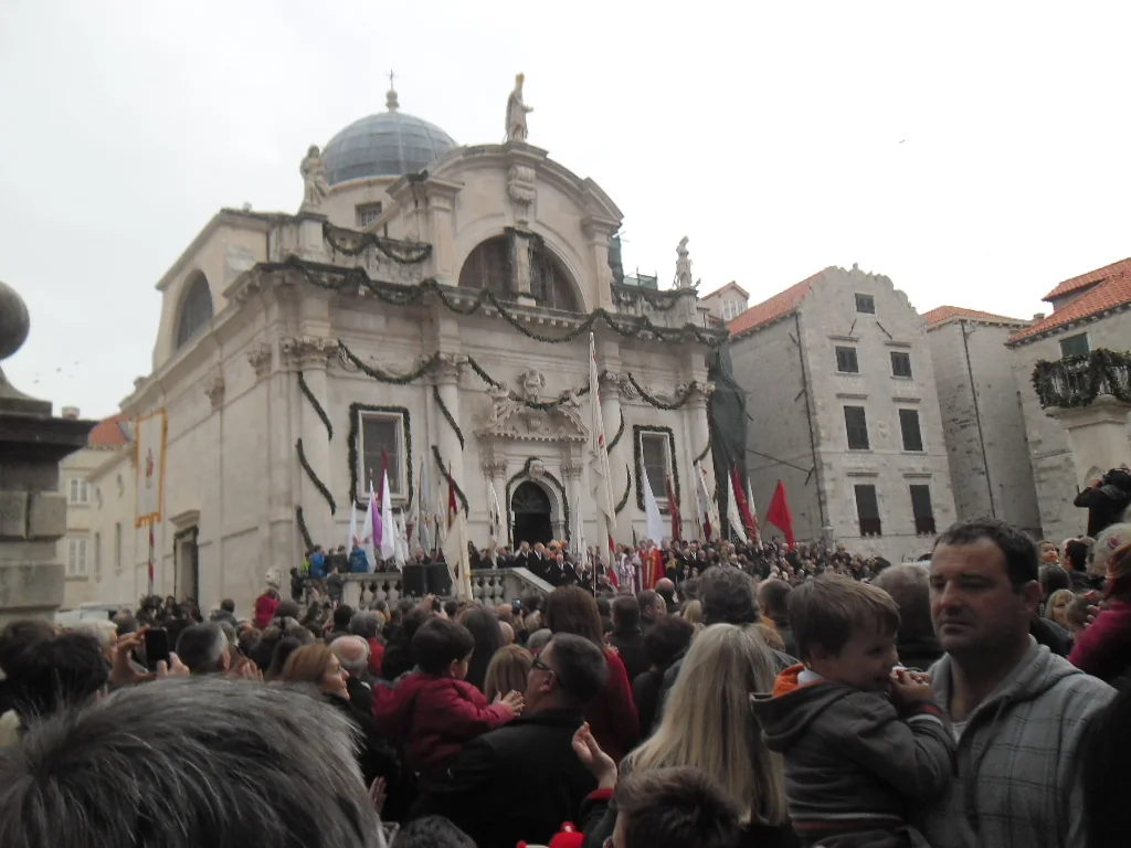 A crowd on the square in front of the Church of St Blaise at the Candlemas opening, with garlands across the facade and flags standing on the church steps