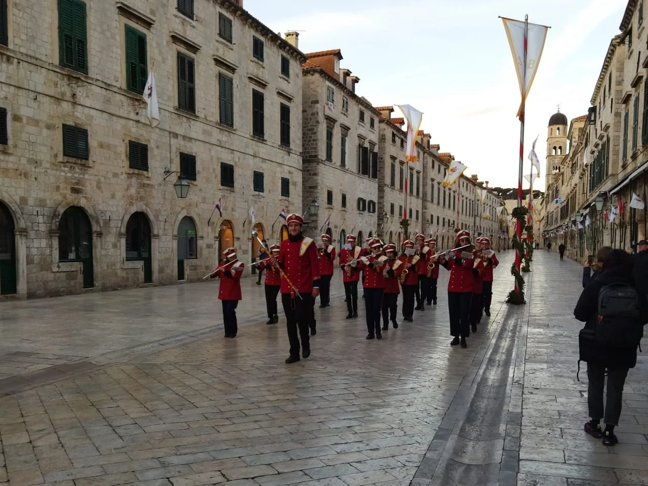 The Dubrovnik city band in red uniforms marching down an almost empty Stradun between poles wound with laurel and hung with white banners of the saint