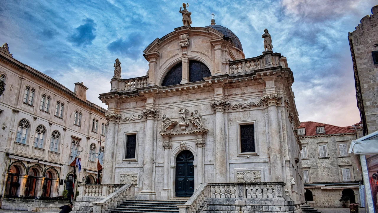 The Baroque facade of the Church of St Blaise in Dubrovnik at dusk, with its flight of steps, the saint's statue on the roof and festive garlands on the building to the left