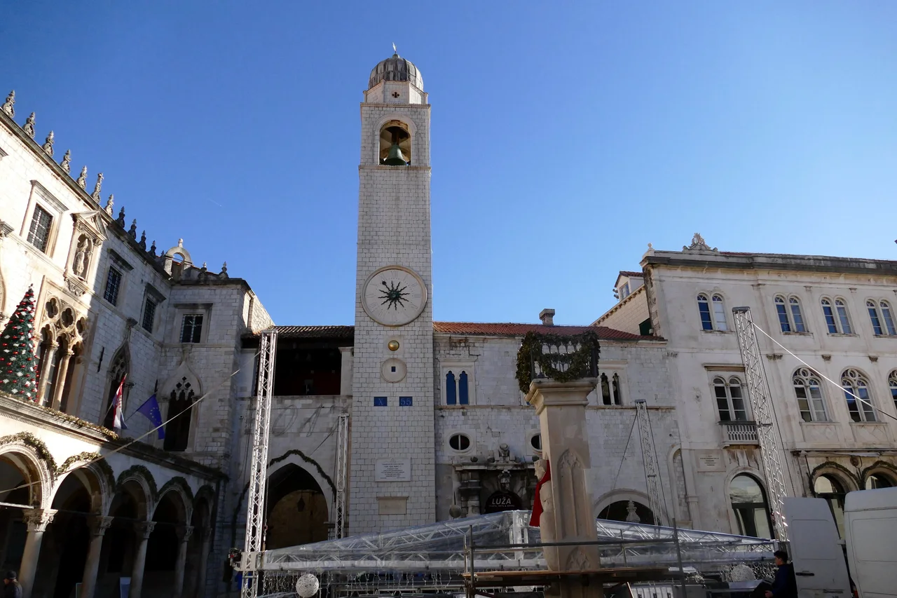 Luza Square in winter sun: the Bell Tower with its clock, the Gothic arcade of the Sponza Palace, a Christmas tree and Orlando's Column wearing a red scarf, with a stage going up in front