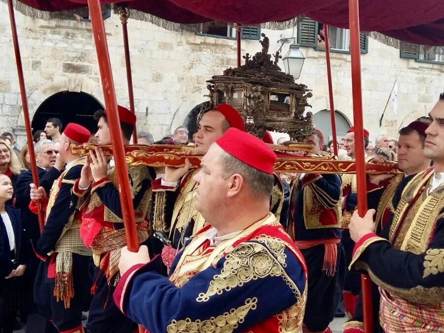 Bearers in red caps and embroidered folk costume carrying a gilded reliquary under a crimson canopy through a stone-paved street