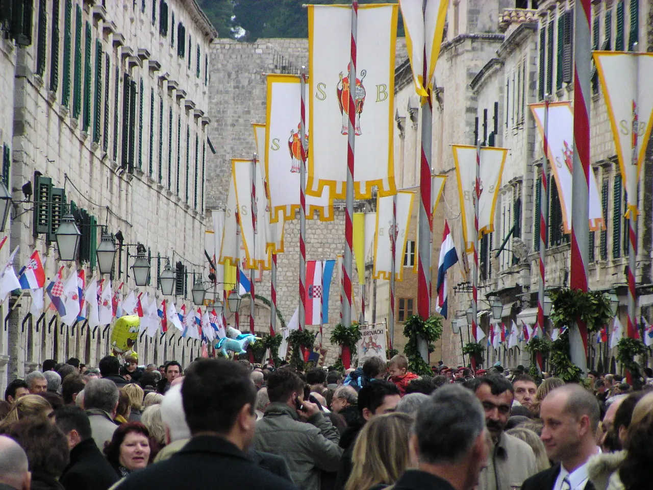 Stradun packed with people and hung with tall white banners of St Blaise on striped poles, with rows of small flags along the facades