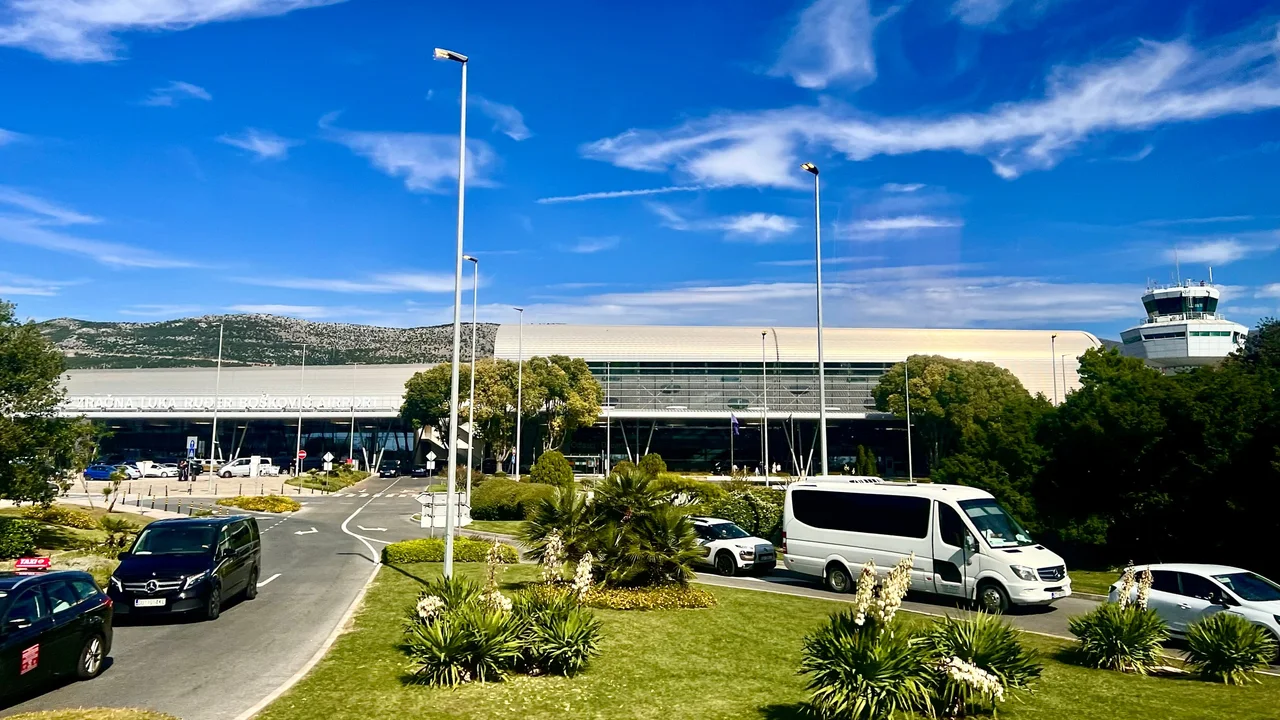 Dubrovnik Airport's terminal with its long curved roof, taxis and a minibus at the kerb, the control tower on the right and a rocky hill behind