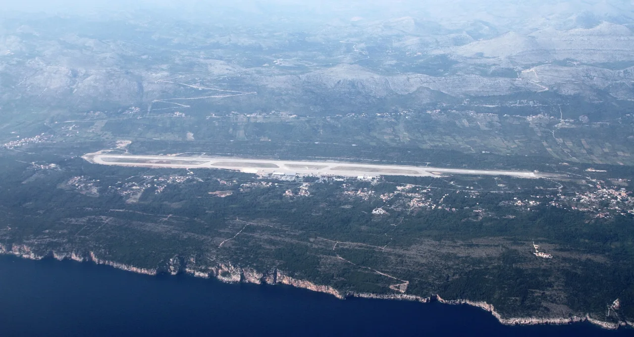 Aerial view of Dubrovnik Airport's runway on the Konavle coast, with the dark Adriatic and a line of limestone cliffs below it and hazy hills behind