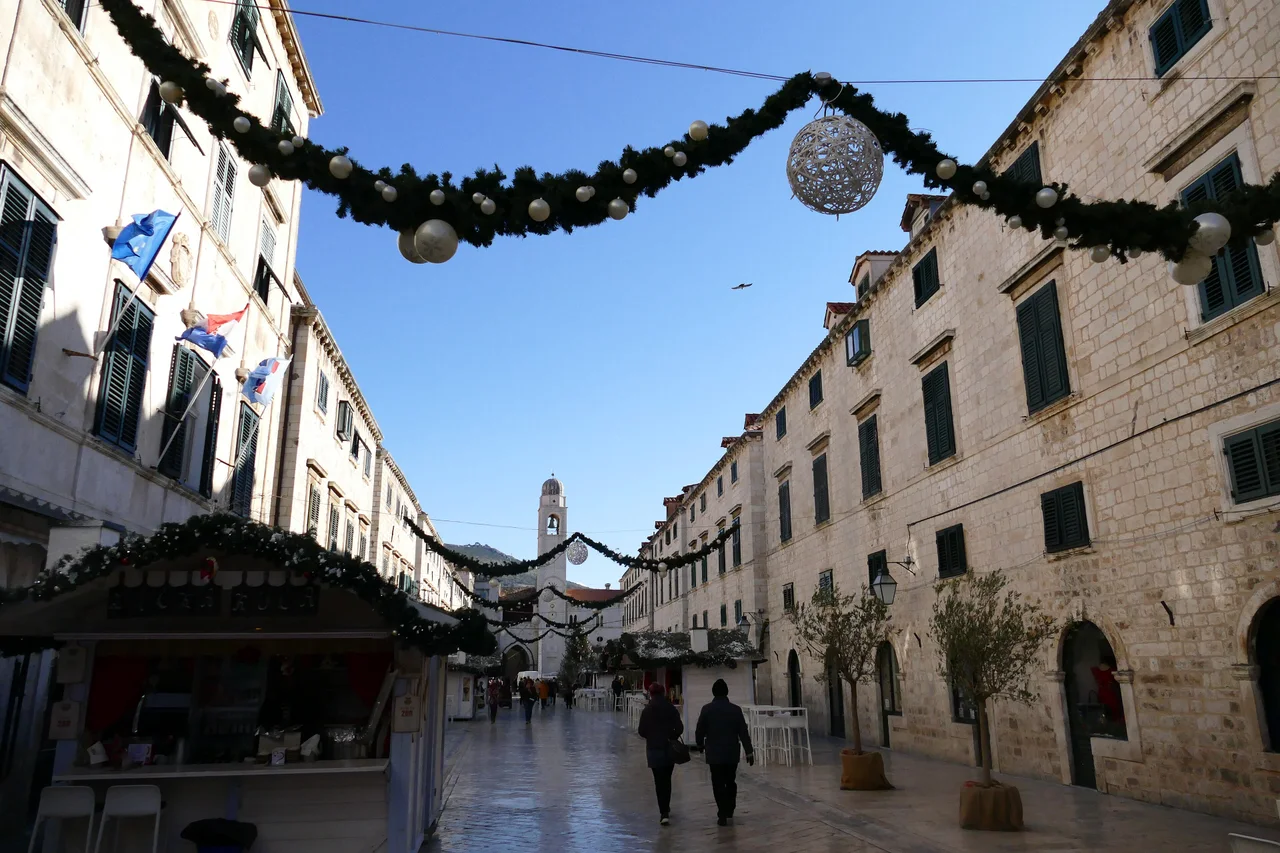 Stradun, the main street of Dubrovnik's Old Town, in winter sun with Christmas garlands and baubles overhead, market stalls on the left and the bell tower at the far end