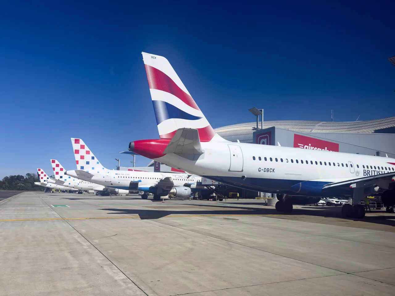 The red, white and blue tail of a British Airways Airbus in the foreground, with a row of Croatia Airlines tails in their red-and-blue checkerboard pattern behind it on the apron at Zagreb Airport