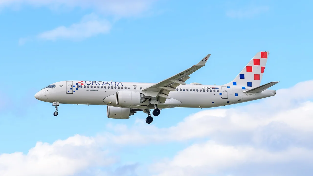 A Croatia Airlines Airbus A220-300 on approach to London Heathrow, landing gear down, white fuselage and red-and-blue checkerboard tail against a blue sky with clouds