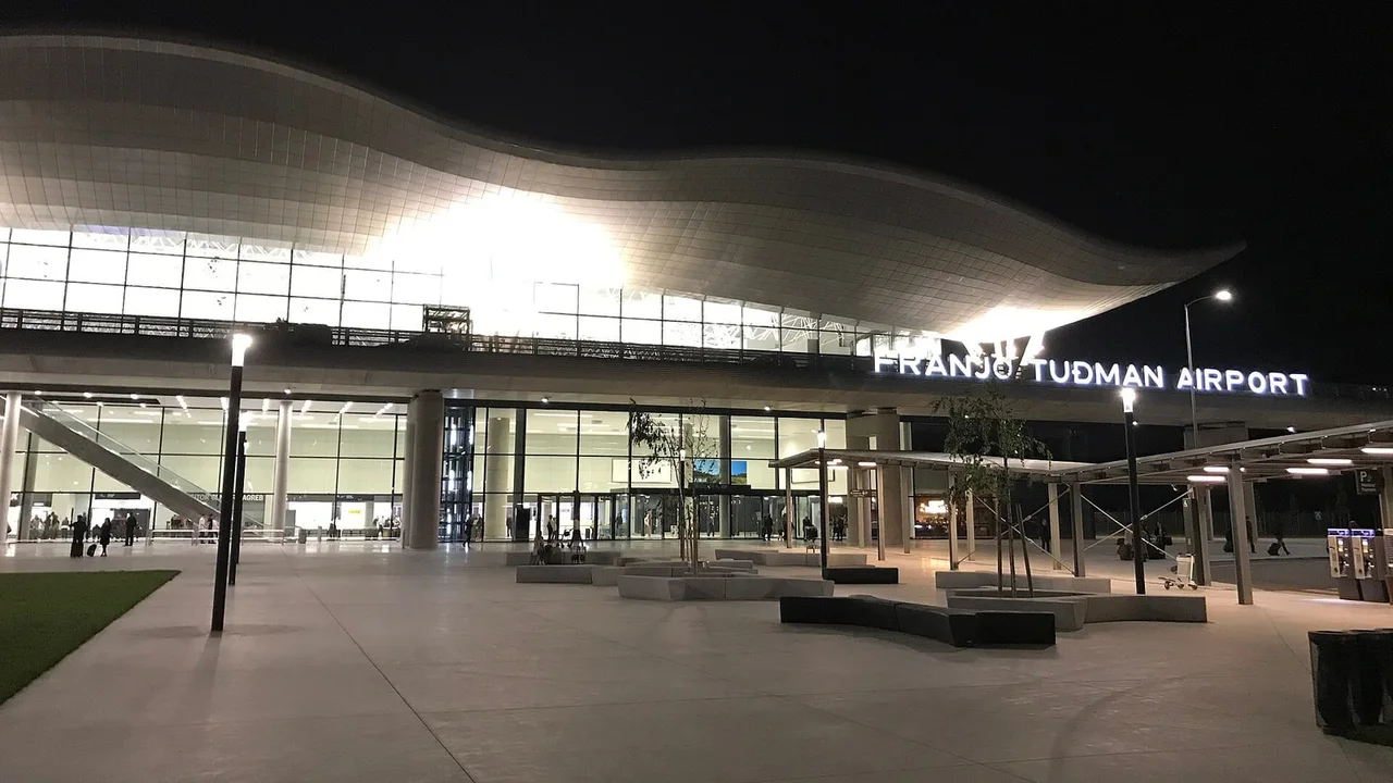 The lit wave-shaped roof and glass front of the Franjo Tuđman Airport terminal at night, with a paved forecourt, benches and young trees in front