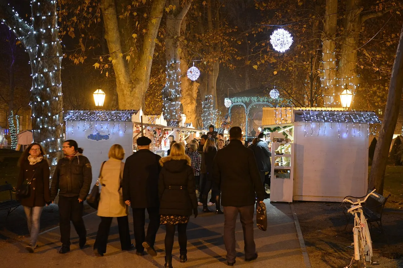 Visitors strolling past white Advent stalls on a path in Zagreb's Zrinjevac park, plane trunks wrapped in white lights, a glowing ball hanging overhead and the lit bandstand behind